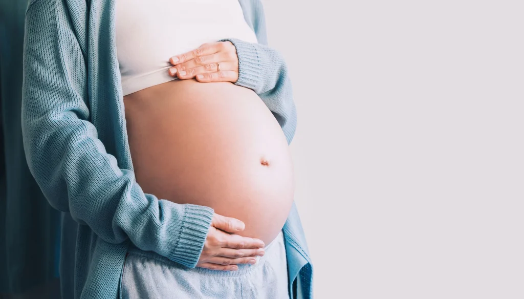 Pregnant woman cradling her belly, wearing a light blue cardigan over a white top, against a neutral background.