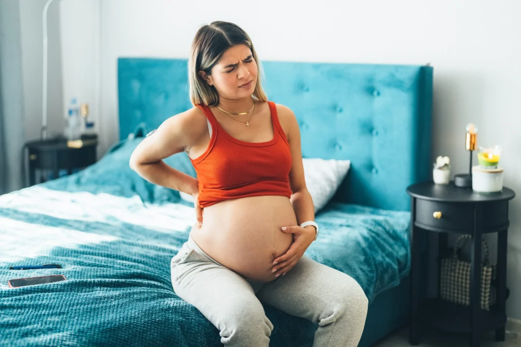 Pregnant woman in a red tank top sitting on a bed, holding her back and appearing uncomfortable.